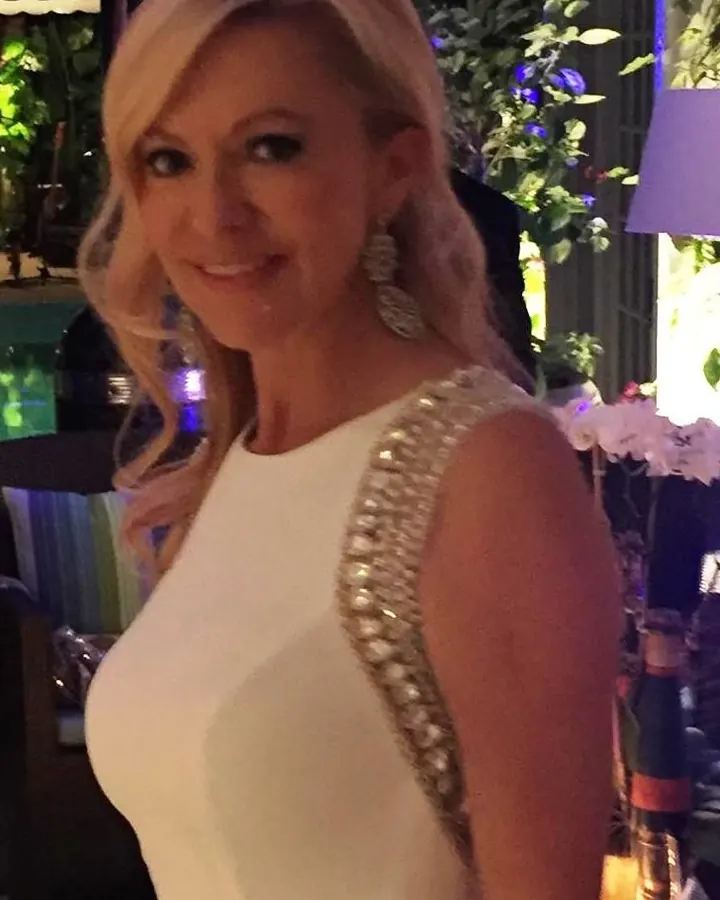 Femme blonde souriante en robe blanche avec strass sur l'épaule, dans un cadre intérieur éclairé par des lumières colorées.