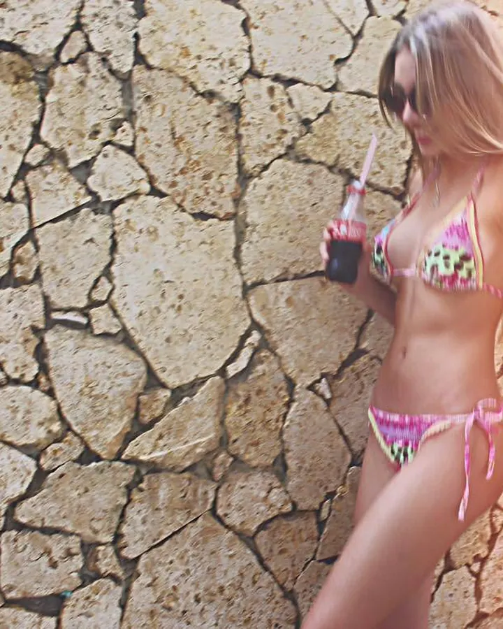 Femme blonde en maillot de bain multicolore, tenant une boisson, appuyée contre un mur en pierre sous un soleil chaud.