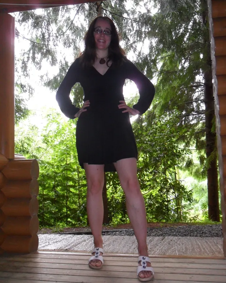 Femme aux cheveux châtains, debout dans un jardin, portant une jupe orange, un haut blanc et une veste noire à boutons dorés, tenant un sac à main marron.