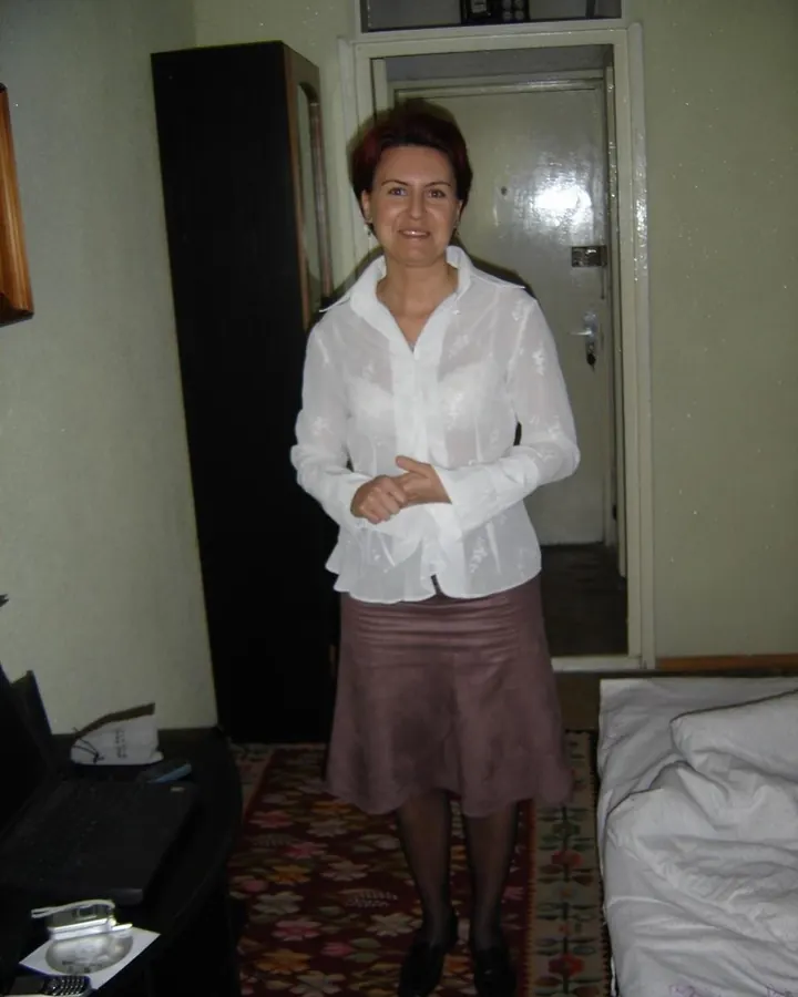 Femme aux cheveux châtains, debout dans une chambre, portant une chemise blanche transparente et une jupe marron, souriant légèrement.