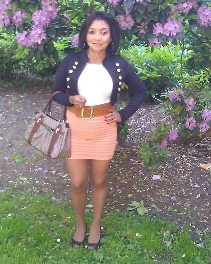 Femme arabe de 45 à 60 ans, debout dans un jardin fleuri, portant une jupe orange, un haut blanc et une veste noire à boutons dorés, tenant un sac à main marron.