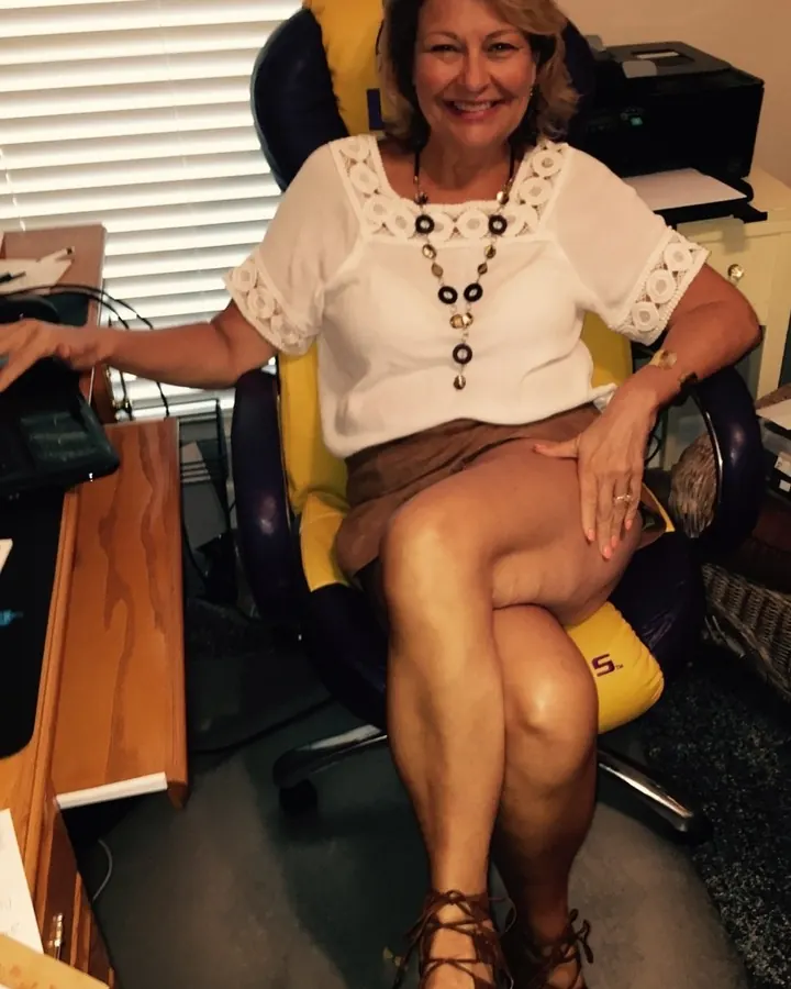 Femme blonde de 45 à 60 ans, assise sur un fauteuil de bureau jaune et bleu, portant une blouse blanche à dentelle, une jupe marron et des sandales à lacets.