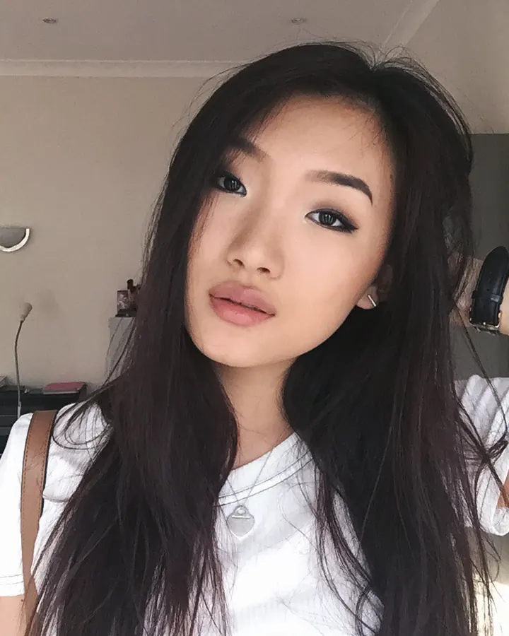Femme asiatique aux cheveux bruns noirs, portant un t-shirt blanc, regardant l'appareil photo avec un léger sourire, une main dans ses cheveux.