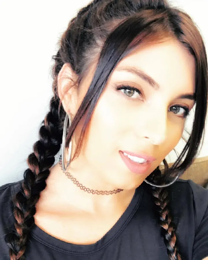 Femme aux cheveux châtains en tresses, souriante, portant un collier fin et des boucles d'oreilles fines, vêtue d'un t-shirt noir.