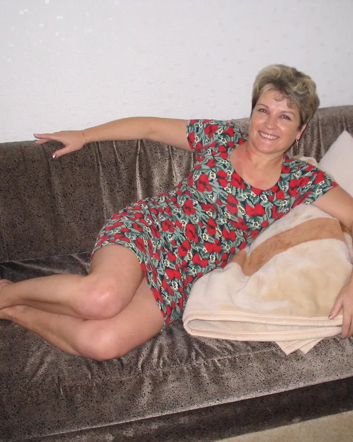 Femme aux cheveux courts blonds, vêtue d'une robe à motifs floraux rouges et verts, allongée sur un canapé marron avec une couverture beige.