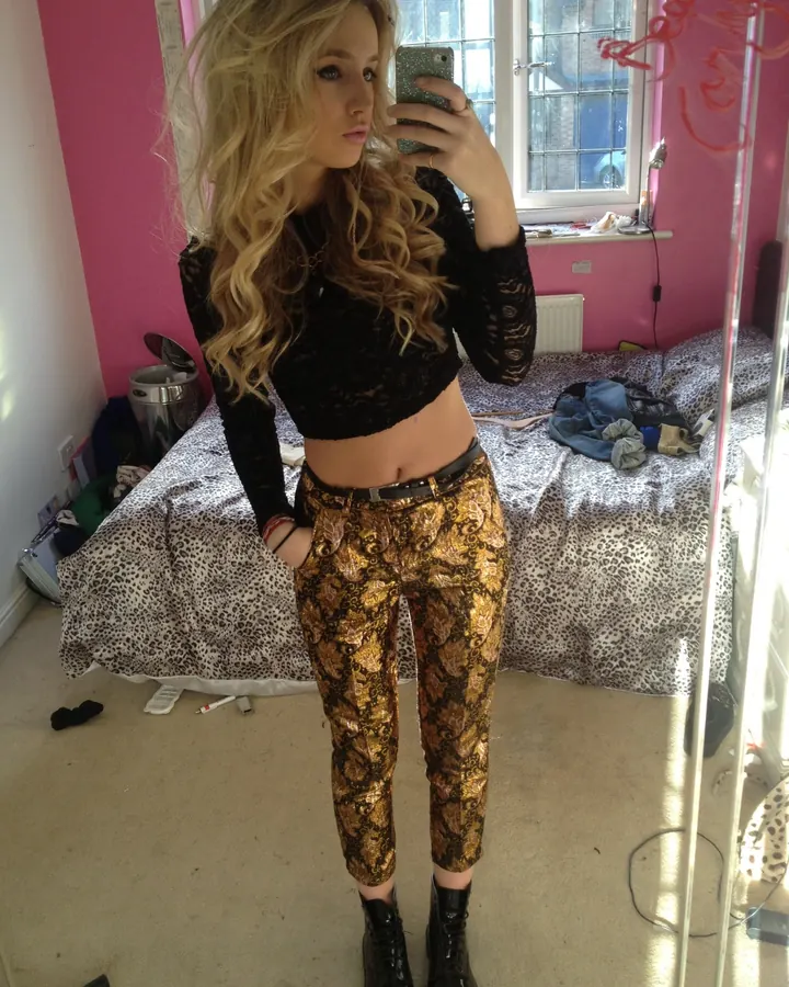 Jeune femme blonde en tenue décontractée (top noir ajouré, pantalon doré à motifs, bottines noires), prenant un selfie devant un miroir dans une chambre aux murs roses.