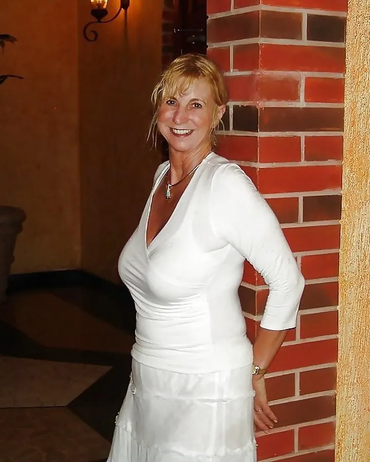 Femme blonde souriante en robe blanche, appuyée contre un mur en briques rouges dans un intérieur chaleureux.
