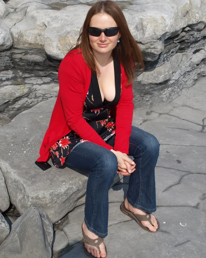 Femme aux cheveux bruns portant une cardigan rouge, un haut noir à fleurs et un jean, assise sur un rocher en extérieur.
