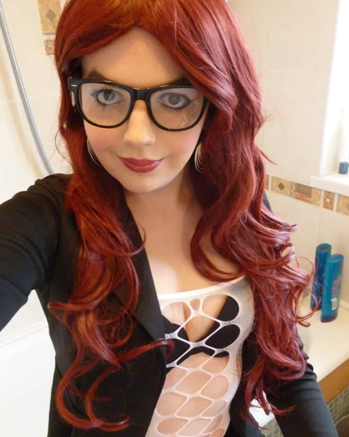 Femme aux cheveux roux portant une perruque, des lunettes noires et un body blanc transparent sous une veste noire, prenant un selfie dans une salle de bain.