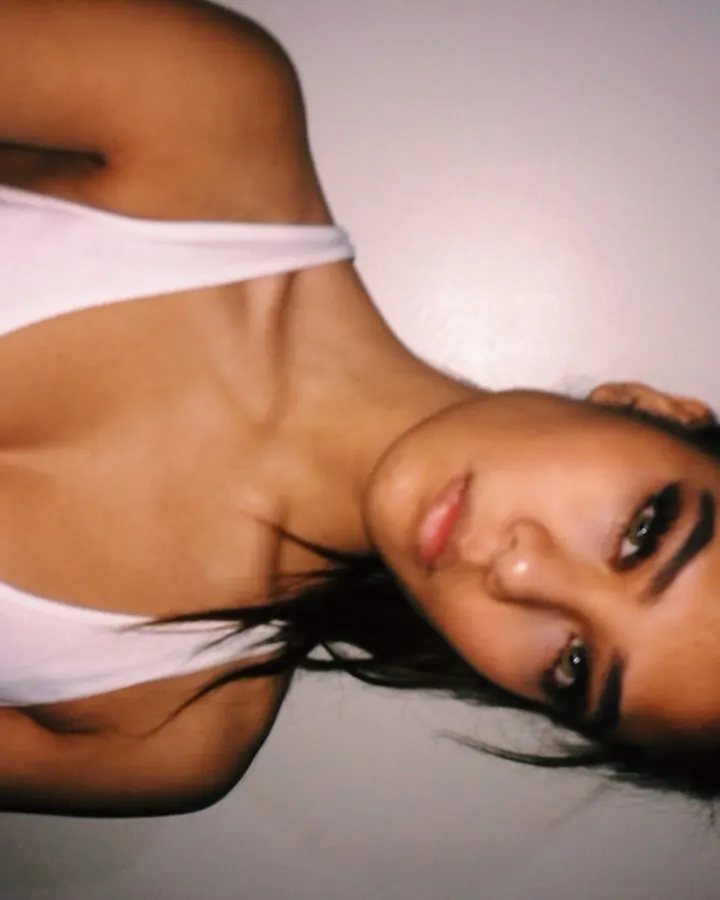 Jeune femme aux cheveux bruns noirs en haut blanc, prenant un selfie devant un lit défaite.
