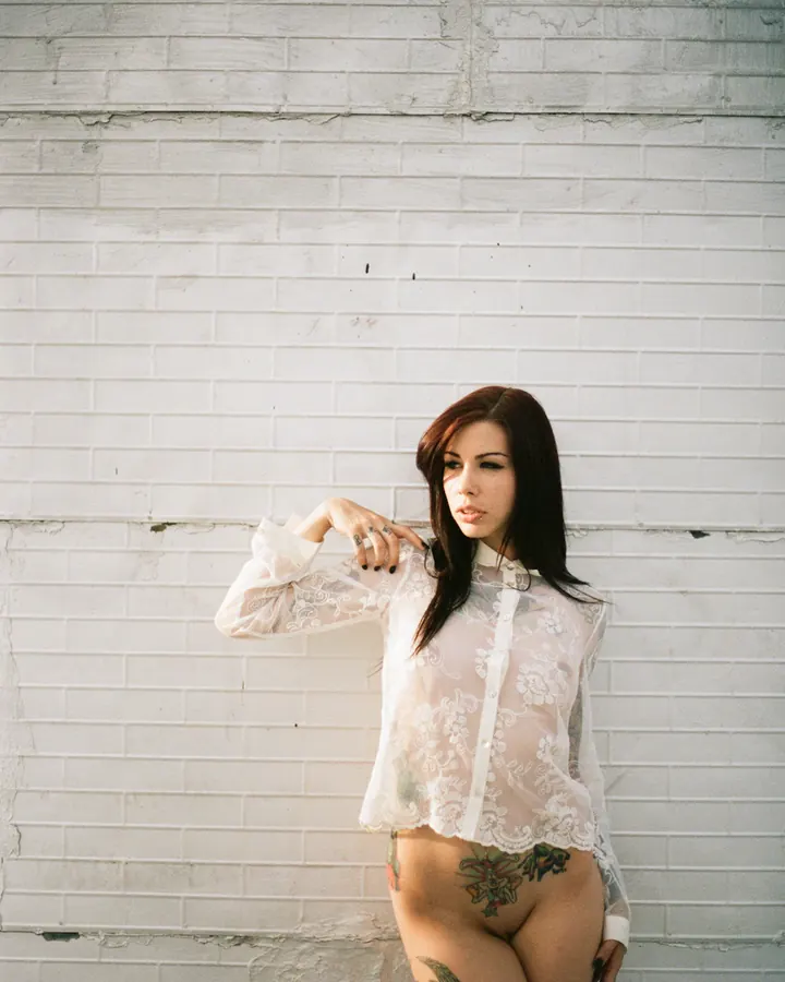 Femme aux cheveux roux, portant une chemise blanche transparente avec dentelle, debout contre un mur de briques blanches, tatouages visibles sur les hanches et les jambes.
