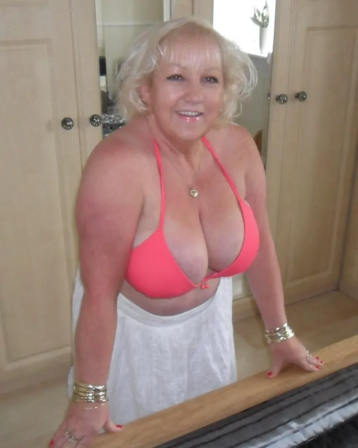 Femme blonde aux yeux bleus, portant un soutien-gorge rose et une jupe blanche, souriant en appui sur un lit.
