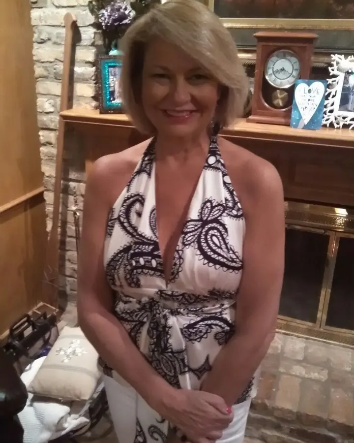 Femme aux cheveux blonds platine en haut à imprimé noir et blanc, souriant à la caméra dans une pièce avec cheminée en pierre.