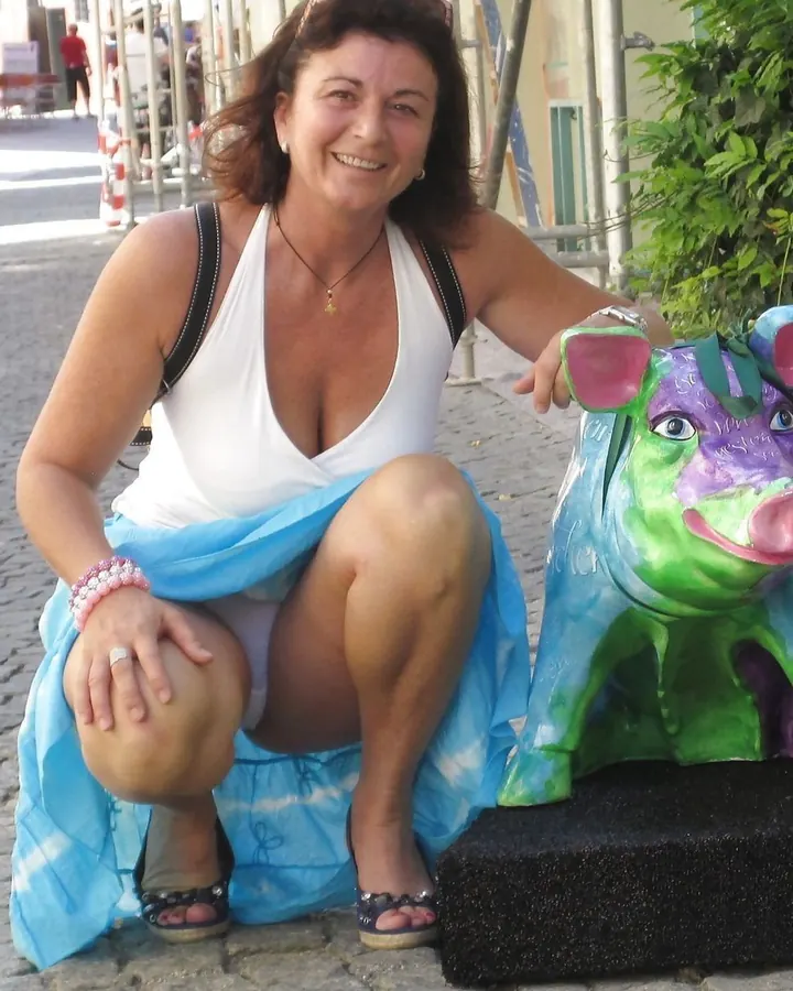 Femme souriante assise sur un trottoir, en haut blanc et jupe bleue, près d'une statue colorée