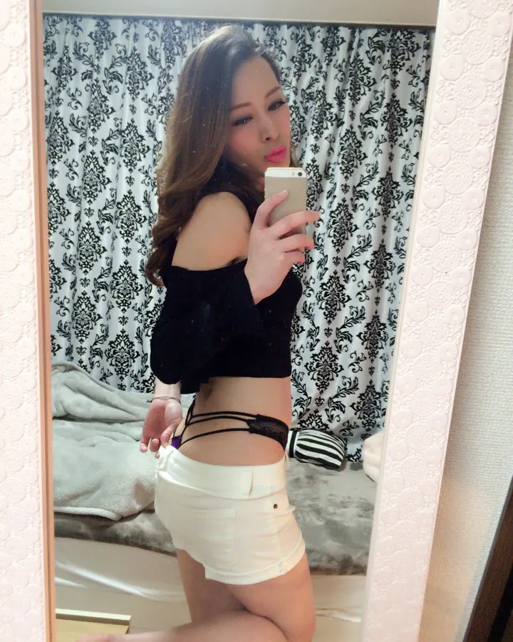 Femme aux cheveux bruns prenant un selfie devant un miroir, en haut noir et short blanc, avec un string noir visible