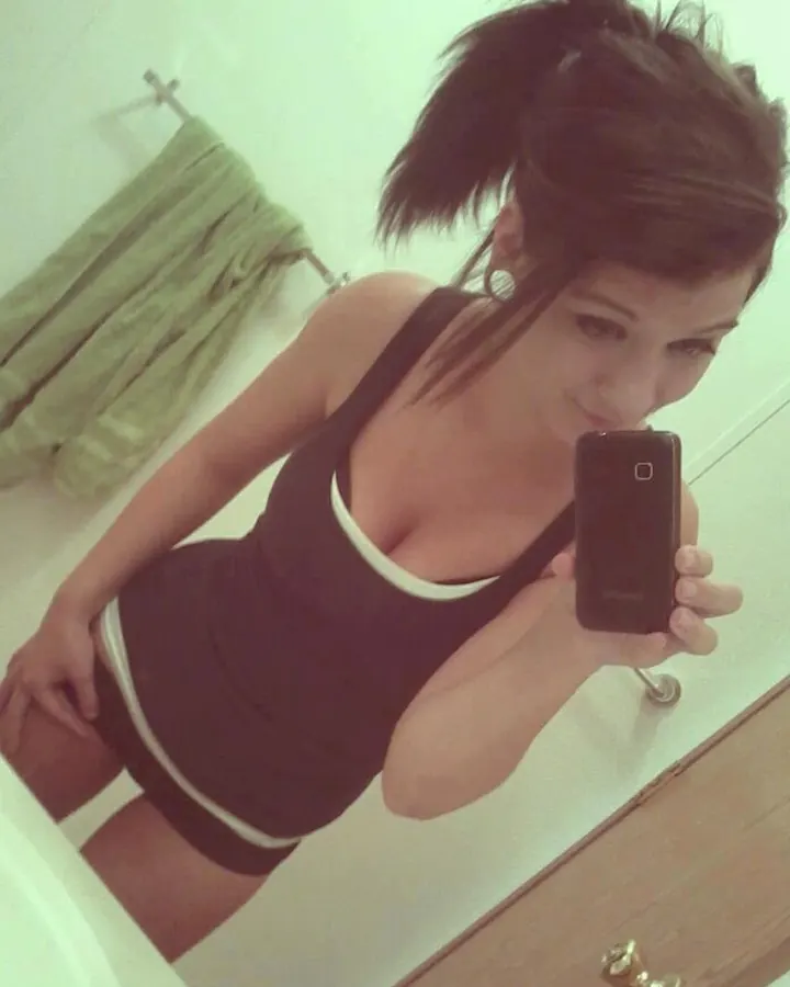 Jeune femme aux cheveux bruns, en haut noir et blanc, prenant un selfie devant un miroir dans une salle de bain.