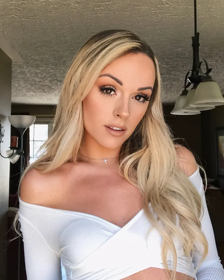 Femme aux cheveux blonds, haut blanc, pose debout près d’un mur, regardant vers la caméra.