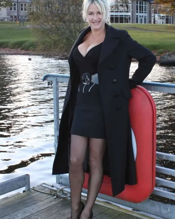 Femme aux cheveux gris, veste noire, jupe noire, collants noirs, chaussures à talons, assise sur un canapé rouge.