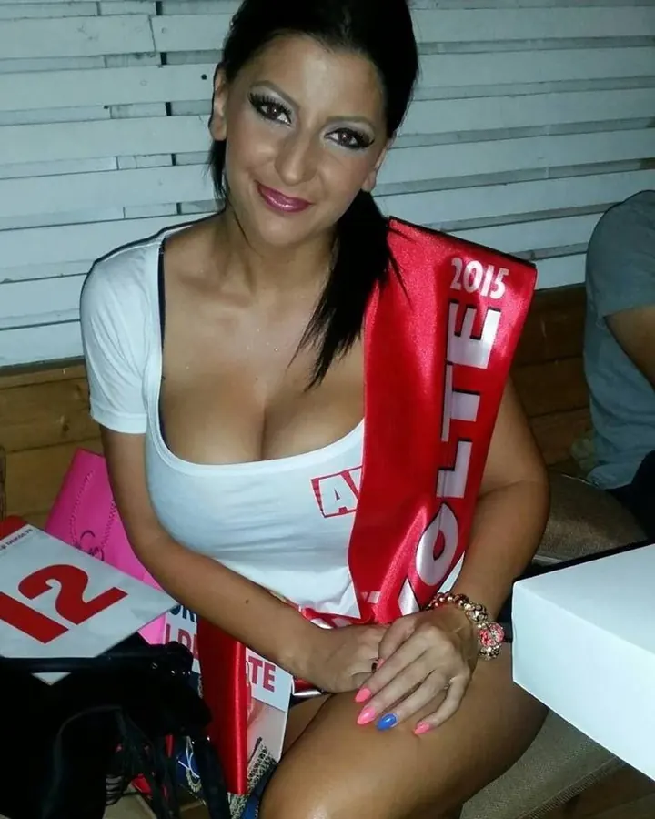 Femme aux cheveux bruns-noirs, maquillée, portant une sash rouge et un t-shirt blanc, assise sur un banc.