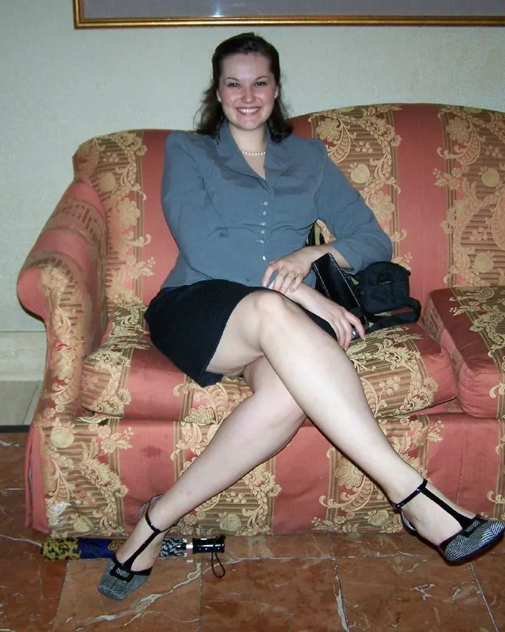 Femme aux cheveux bruns courts portant un haut rose pâle, assise sur un canapé rouge à motifs, les jambes croisées, chaussée de sandales noires à talons, tenant un petit sac noir.