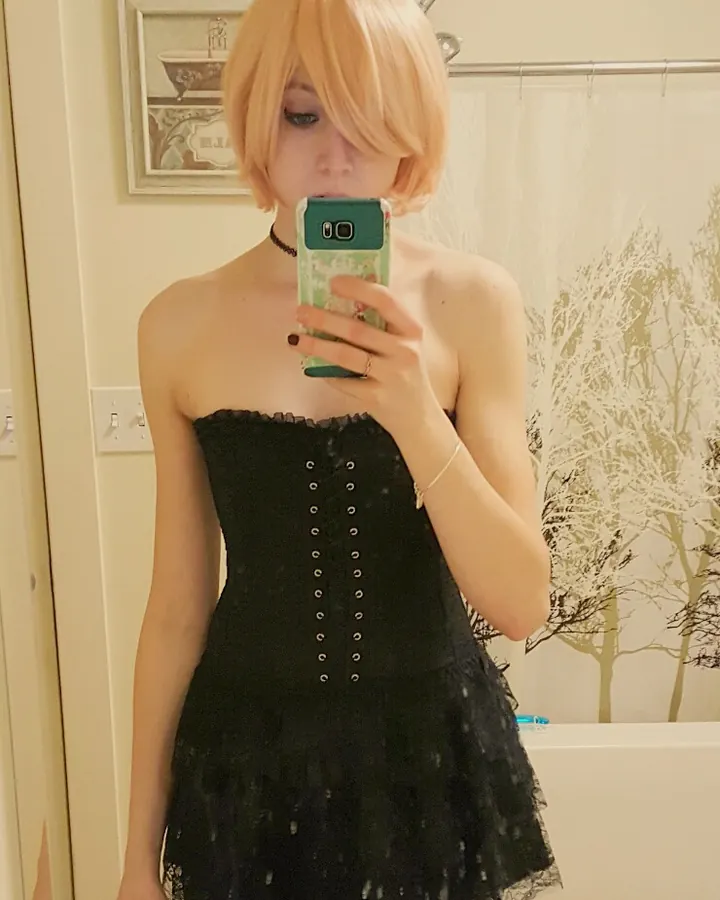 Femme aux cheveux blonds courts prenant un selfie dans un miroir, portant un corset noir et une jupe noire en dentelle.