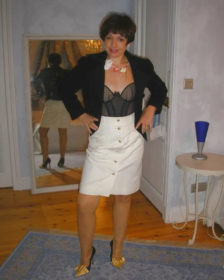 Femme aux cheveux bruns-noirs portant un corset noir sous une veste noire et une jupe blanche boutonnée, debout devant un miroir dans une pièce aux murs clairs.