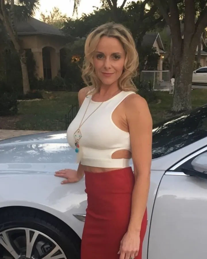 Femme blonde portant un haut blanc et une jupe rouge, appuyée sur une voiture blanche, souriant légèrement, dans un cadre résidentiel.