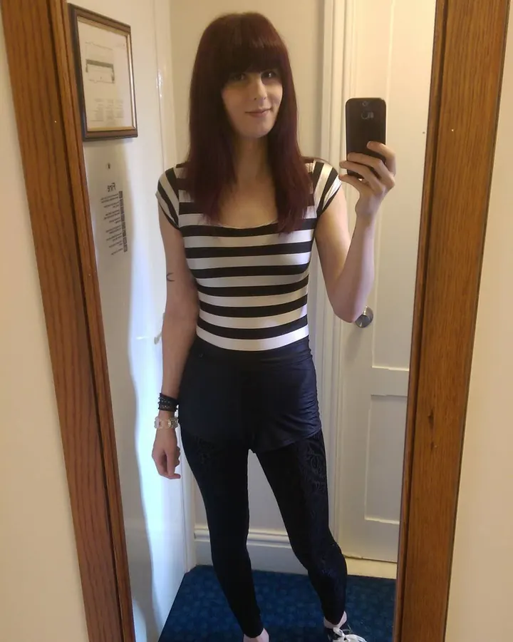 Femme aux cheveux châtains portant un haut noir et un short noir, prenant un selfie dans un miroir, avec des leggings noirs ornés de motifs.