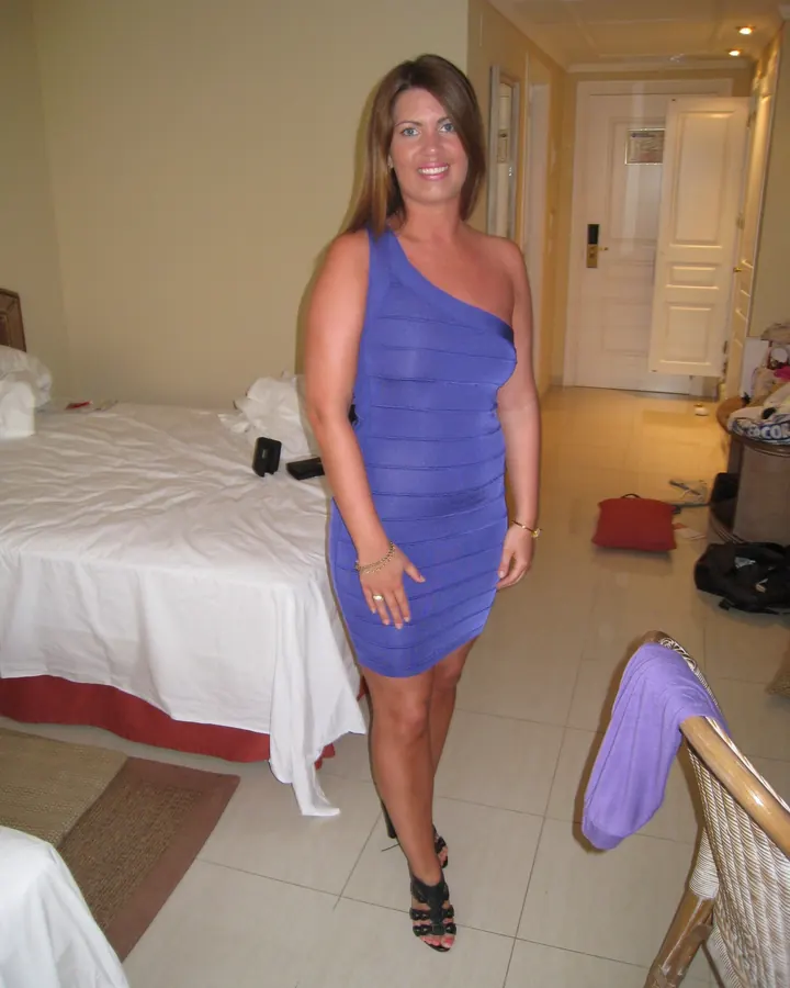 Femme aux cheveux bruns portant une robe bleue ajustée, debout dans une chambre d'hôtel.