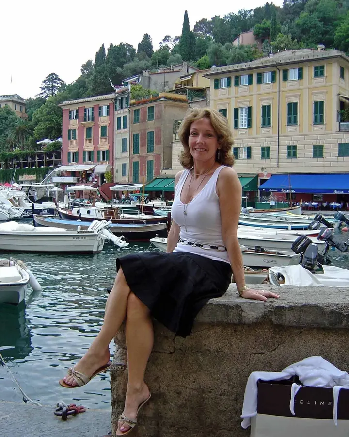 Femme aux cheveux blonds courts, portant un haut blanc et une jupe noire, assise sur un mur de pierre près d'un port avec des bateaux, souriant vers l'objectif.