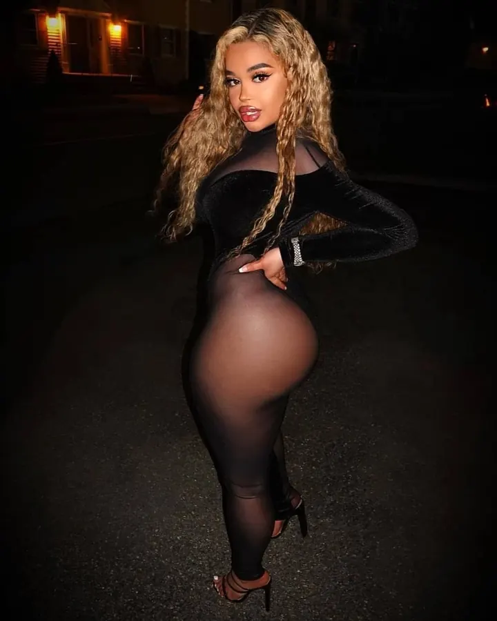 Femme aux cheveux blonds ondulés, portant une tenue noire en velours et maille transparente, posant de profil sur une route la nuit, avec des lumières en arrière-plan.