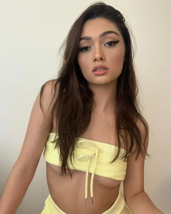Jeune femme asiatique aux yeux bleus et cheveux châtains, portant un haut jaune fluo sans bretelles, regardant l'objectif avec un maquillage soigné, dans un intérieur clair.