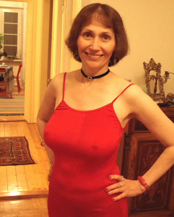 Femme aux cheveux bruns courts, portant une robe rouge ajustée, souriant, une main sur la hanche, dans un intérieur chaleureux avec un meuble en bois et des bougies rouges en arrière-plan.