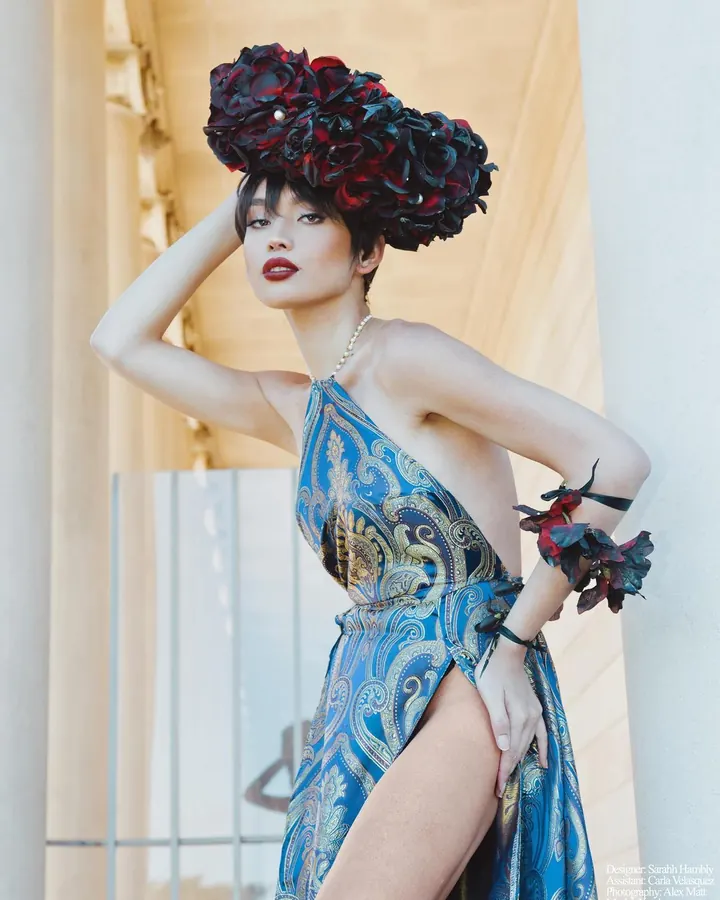 Femme asiatique en robe bleue à motifs dorés, coiffée d'un chapeau de fleurs rouges et noires, posant près d'une colonne.