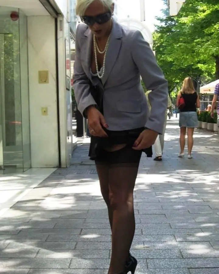 Femme aux cheveux blancs en costume gris, lunettes de soleil, tenant sa jupe courte sur un trottoir ombragé.