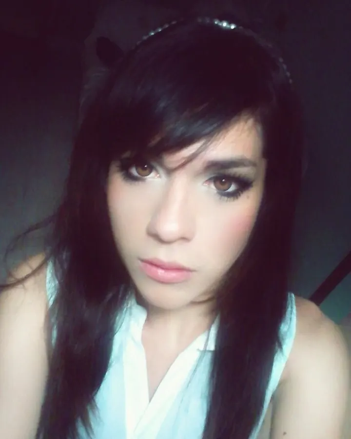 Femme aux cheveux noirs, maquillage prononcé, regardant l'appareil photo dans un cadre sombre, portant un haut blanc.