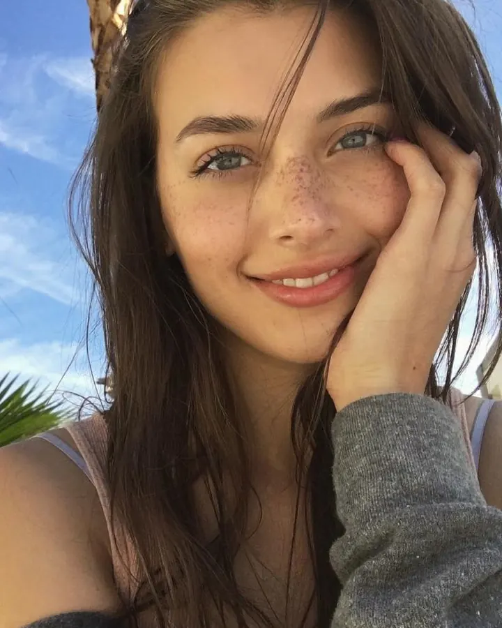 Femme en haut rose, souriant, main sur le visage, en extérieur.