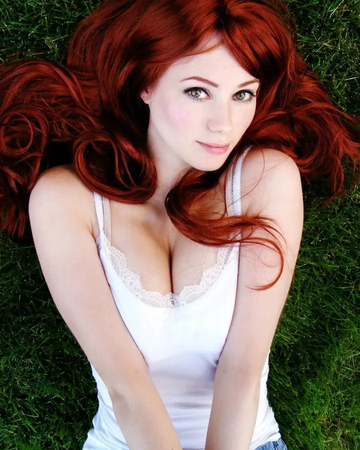 Femme aux cheveux roux allongée sur l'herbe, regardant vers l'appareil, sourire discret.