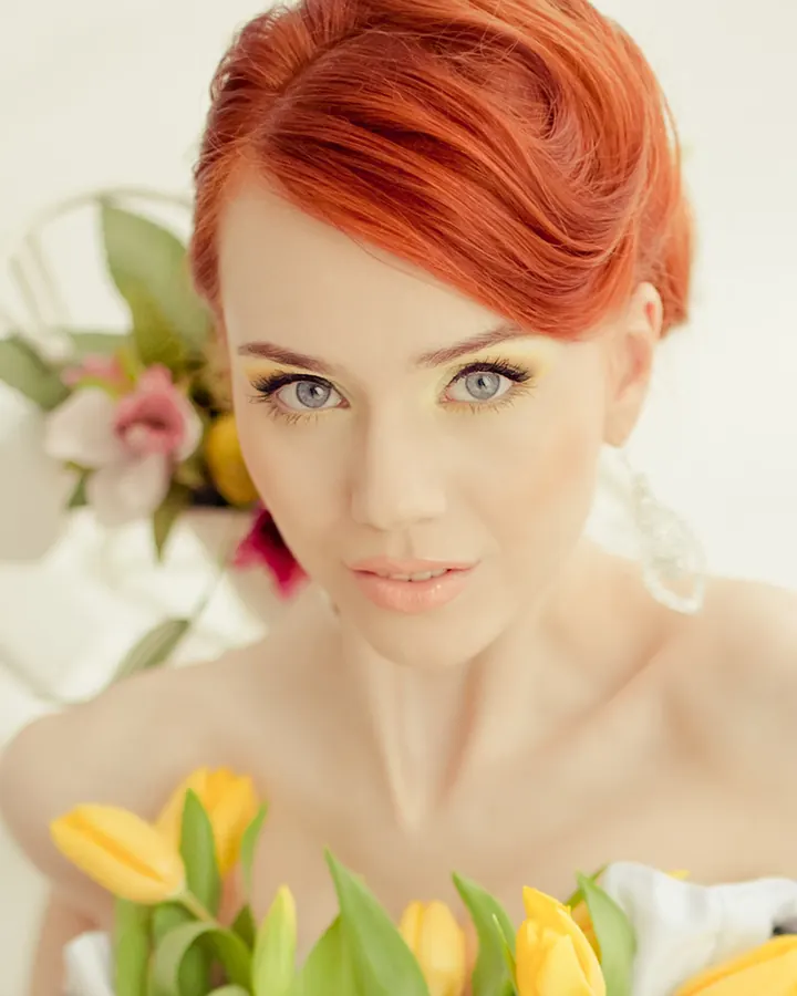 Femme aux cheveux roux, maquillée, tenant des tulipes jaunes, avec un fond blanc et fleuri.