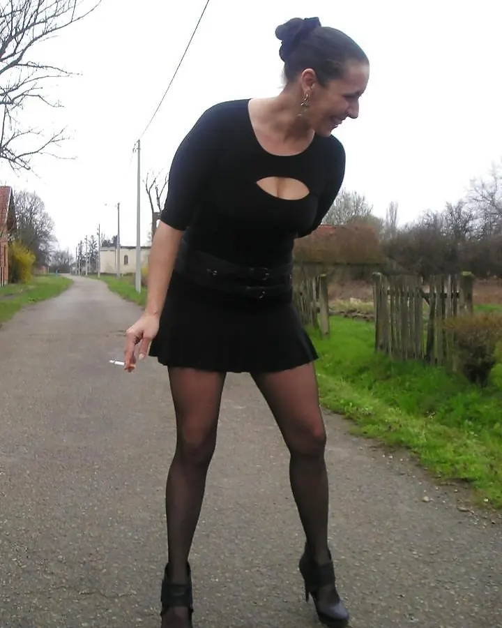 Femme aux cheveux bruns foncés, vêtue d’un haut noir et d’une jupe noire courte, portant des collants à mailles et des chaussures à talons, posant dans une cuisine.