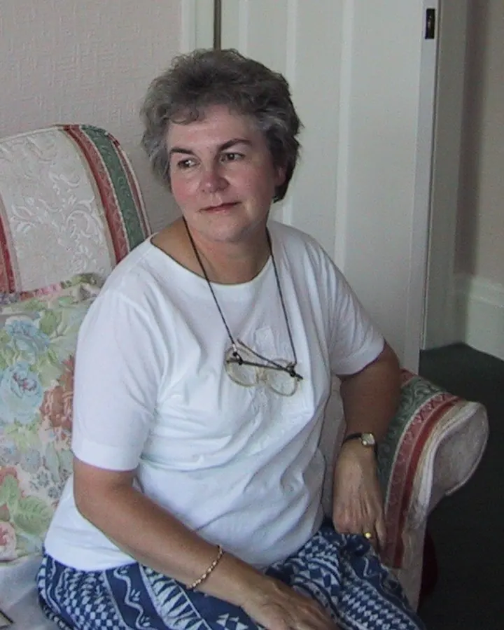Femme aux cheveux gris, souriante, portant un haut rose, assise sur un banc, dans un jardin verdoyant.