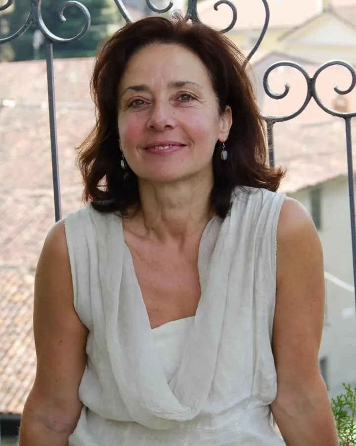 Femme aux cheveux bruns, en tenue blanche, assise sur une chaise métallique devant une fenêtre, souriante.