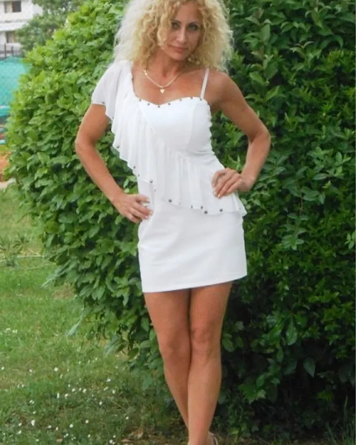 Femme aux cheveux blonds bouclés en robe blanche, posant devant un buisson vert