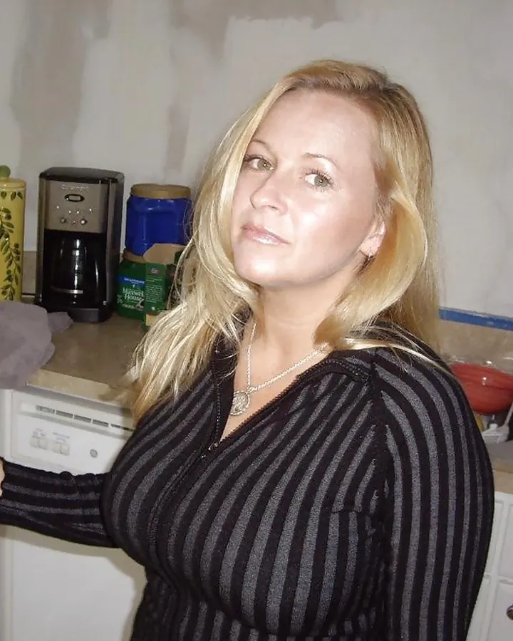 Femme européenne aux cheveux platine, portant un haut noir décolleté, posant dans une cuisine, souriant légèrement.