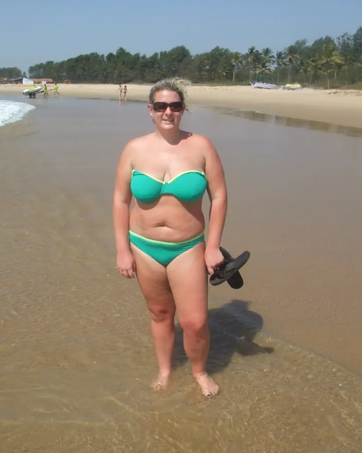 Femme européenne aux cheveux bruns-noirs, portant un haut blanc sans manches, debout dans une cuisine, souriant largement.