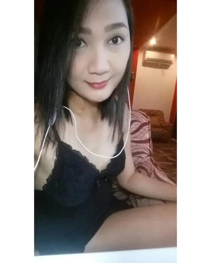 Femme asiatique aux cheveux bruns-noirs, portant un haut noir en dentelle, assise sur un lit dans une chambre, avec des écouteurs blancs.