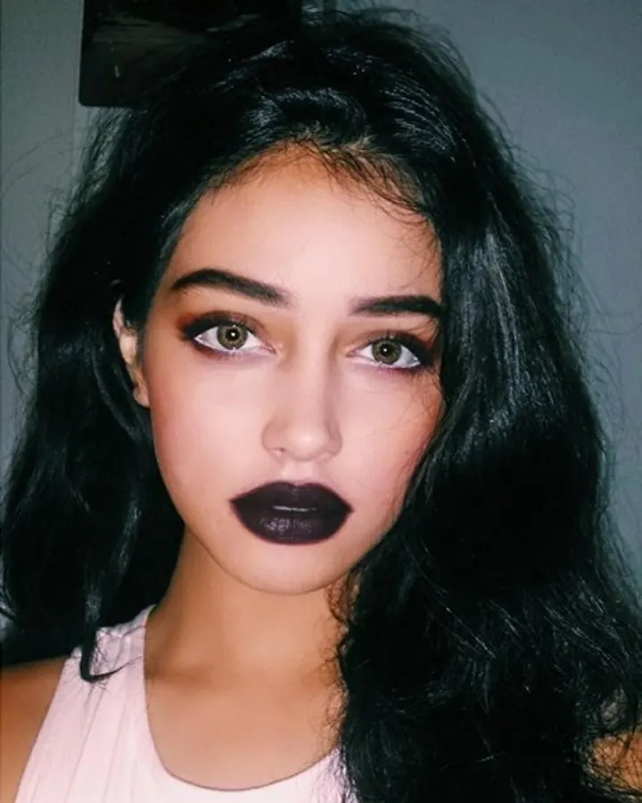 Jeune femme aux cheveux foncés, portant un haut rose pâle, avec un maquillage sombre aux yeux et aux lèvres, regardant directement l'appareil photo.