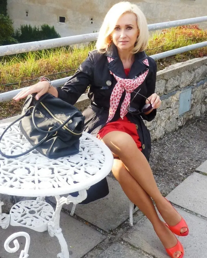 Femme aux cheveux blonds, portant un haut noir et une jupe noire, assise sur une chaise dans une cuisine, avec une table devant elle ayant des tasses et des objets divers.
