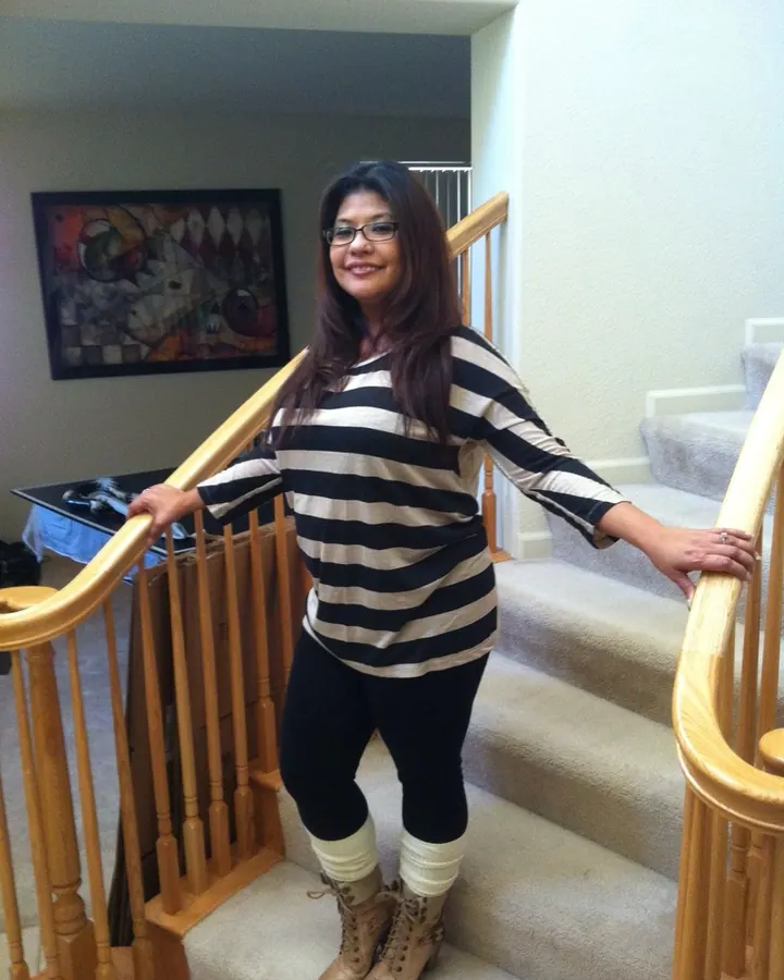 Femme aux cheveux longs bruns, portant un haut rayé noir et beige, pantalon noir, bottines marron et bas blancs, souriante en se tenant à une rampe d'escalier intérieur.