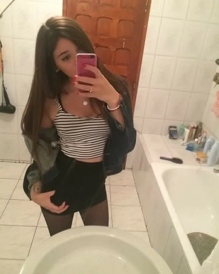 Jeune femme aux cheveux bruns prenant un selfie dans une salle de bain, vêtue d'un haut rayé noir et blanc, d'une jupe noire courte et de collants noirs, avec une veste en jean ouverte.